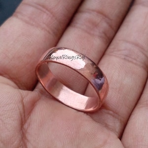 Puede incluir: Un anillo de color cobre con una banda lisa y redondeada. El anillo tiene un acabado pulido y el texto "RoyalRingsRaj" es visible en la superficie. El anillo es de diseño simple y clásico.