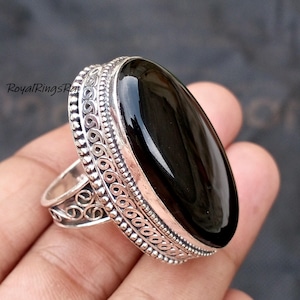 May include: A large, oval-shaped black gemstone ring with an ornate silver band. The ring features intricate detailing around the stone and along the sides of the band. The gemstone is polished and reflects light.
