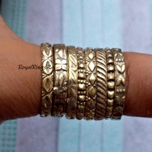 May include: A collection of gold-colored rings with various embossed patterns, including floral, geometric, and textured designs. The rings are stacked on a finger, showcasing the different styles and textures. The rings are likely made of metal and are suitable for everyday wear.