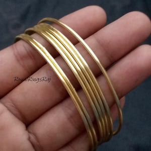 May include: A stack of thin, gold-colored bangles. The bangles are smooth and shiny, reflecting light. The image is taken from a close-up angle, showing the details of the jewelry. The text "Royal Rings Raj" is visible in the lower left corner.