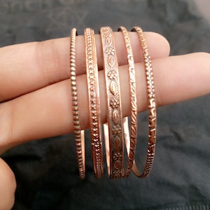 Peut inclure: Ensemble de cinq bracelets de couleur cuivre, chacun avec un design unique. Les bracelets présentent différentes textures et motifs, notamment des perles, des motifs floraux et géométriques. Les bracelets sont d'une couleur brun-rougeâtre chaude.