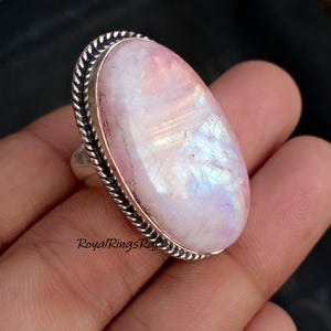 May include: A large oval moonstone ring with a silver band and a textured border. The moonstone has a pearlescent sheen with pink and blue hues. The ring is a close-up shot, showcasing the details of the gemstone.
