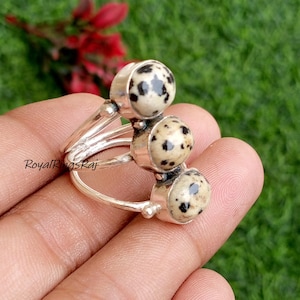 Może przedstawiać: Srebrny pierścionek z trzema okrągłymi, nakrapianymi kamieniami szlachetnymi. Kamienie szlachetne są kremowe z czarnymi plamkami, osadzone w srebrnych oprawach. Pierścionek ma podzieloną konstrukcję trzonu.