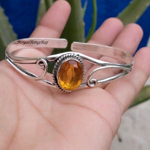 Könnte beinhalten: Ein offener Armreif mit einem zentralen, oval geschliffenen gelben Citrin, der in einer gedrehten Silberfassung sitzt. Das Armband besteht aus poliertem Silber mit dekorativen Drahtwirbeln an beiden Seiten und dem Text RoyalRingRaj.