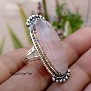 Könnte beinhalten: Ein Silberring mit einem großen, ovalen Rosenquarz-Edelstein. Der Edelstein hat eine zarte rosa Farbe und ist in einem dekorativen Silberband gefasst. Der Ring hat einen Vintage- oder Antik-Stil.