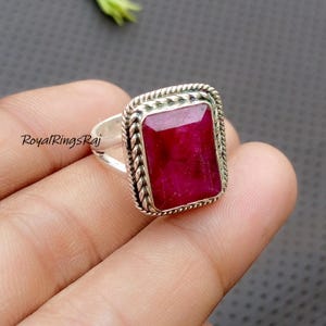 以下が含まれることがあります： 大きな長方形の深い赤色の宝石が付いたシルバーリング。 宝石は装飾的なロープのような縁で囲まれています。 リングは人が持っています。