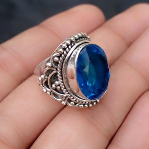 May include: A silver ring with an oval, faceted, bright blue gemstone. The ring has an ornate design with beaded and twisted rope detailing around the gemstone and band. The ring is set against a neutral background.