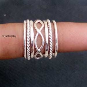 Könnte beinhalten: Ein Satz von fünf Silberringen, die auf einem Finger übereinander liegen. Die Ringe sind in verschiedenen Stilen, darunter ein Seil-Design, ein schlichtes Band und ein Unendlichkeitszeichen.