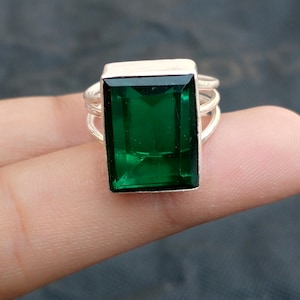 May include: A silver ring with a large, rectangular emerald-green gemstone. The ring has a simple design with a band that splits into multiple strands on each side of the gemstone. The gemstone is the focal point, showcasing its vibrant colour and clarity.