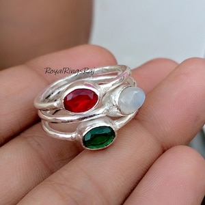 May include: Three silver rings stacked together, each with an oval gemstone: a red ruby, a white moonstone, and a green emerald. The rings are displayed on a hand, with the text "RoyalRingsRaj" visible.