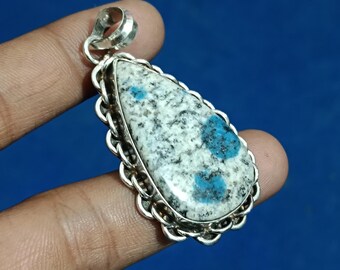 Könnte beinhalten: Tropfenförmiger Anhänger mit einem K2-Stein in einem silberfarbenen Metallrahmen. Der Stein hat ein weißes und graues gesprenkeltes Muster mit blauen Flecken. Der Anhänger hat eine dekorative kettenartige Bordüre und eine Schlaufe für eine Kette.