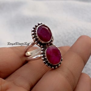 Op de afbeelding: Een zilveren ring met twee ovale, dieprode edelstenen. De edelstenen zijn gezet in een kralenrandontwerp. De ring heeft een gespleten schacht. De tekst "RoyalRingsRaj" is zichtbaar.