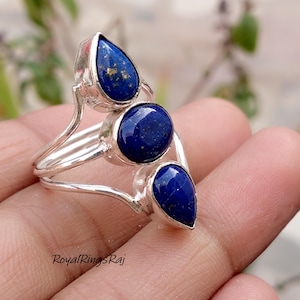 May include: A silver ring featuring three deep blue lapis lazuli stones. The ring has a unique design with a teardrop-shaped stone at the top and bottom, and a round stone in the center. The ring is held in a hand, with the text "RoyalRingsRaj" visible.