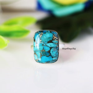 Pode incluir: Um anel retangular de turquesa com uma banda prateada. A pedra turquesa tem uma cor azul-esverdeada vibrante com veios dourados. O anel está sobre um fundo desfocado com tons verdes e azuis. O texto "RoyalRingsRaj" é visível.