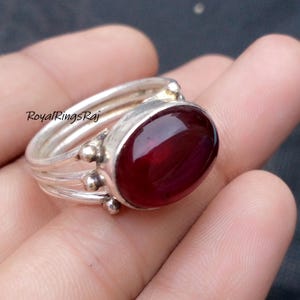 May include: Silver ring featuring a large, oval, deep red gemstone. The ring has a triple band design with small decorative accents. The text "RoyalRingsRaj" is visible on the ring.