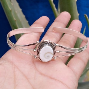 Puede incluir: Una pulsera tipo brazalete de tono plateado con una incrustación central de concha blanca de forma ovalada con un patrón en espiral. La concha está enmarcada por un borde de metal trenzado y montada en una banda de tres hilos metálicos finos.
