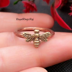 May include: A bronze-colored ring featuring a detailed bee design. The bee has outstretched wings and a segmented body. The ring band is a simple, smooth band. The text "RoyalRingsRaj" is visible in the image.