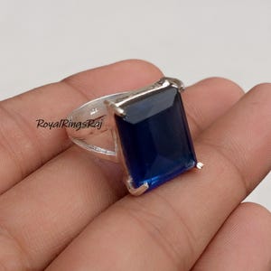 Puede incluir: Un anillo de plata con una gran gema rectangular de color azul oscuro. El anillo tiene una banda sencilla, y la gema está engastada en un marco cuadrado. El texto "RoyalRingsRaj" es visible en la banda del anillo.