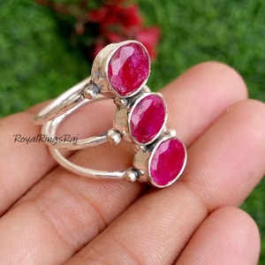 May include: A silver ring featuring three oval-cut, vibrant red gemstones. The ring's band splits into two, with each section supporting a gemstone. The gemstones are set in a bezel setting, and the ring is displayed on a hand.