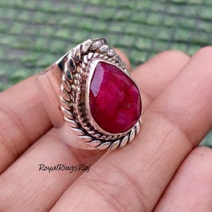 May include: A silver ring featuring a large, teardrop-shaped, faceted ruby gemstone. The ruby is a deep, vibrant red. The ring has a detailed, textured silver band with a braided design around the gemstone. The text "RoyalRingsRaj" is visible.