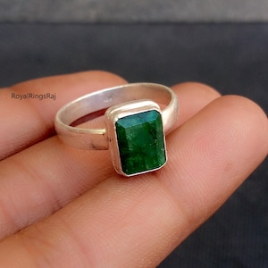 May include: A silver ring with a rectangular, emerald-cut green gemstone. The ring has a simple, polished band and a bezel setting that holds the vibrant green stone. The ring is displayed on a hand.