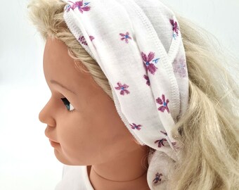 Banda para el cabello para atarse hecha de muselina orgánica con flores dispersas de color púrpura sobre un fondo blanco.