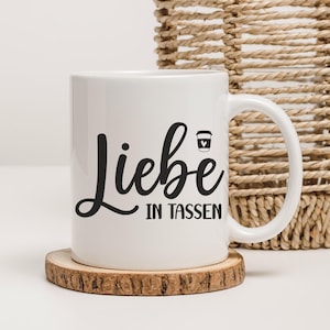 May include: White ceramic mug with the German phrase "Liebe in Tassen" in black script, meaning "Love in Cups." A small coffee cup icon is next to the text. The mug sits on a wooden coaster, with a woven basket in the background.