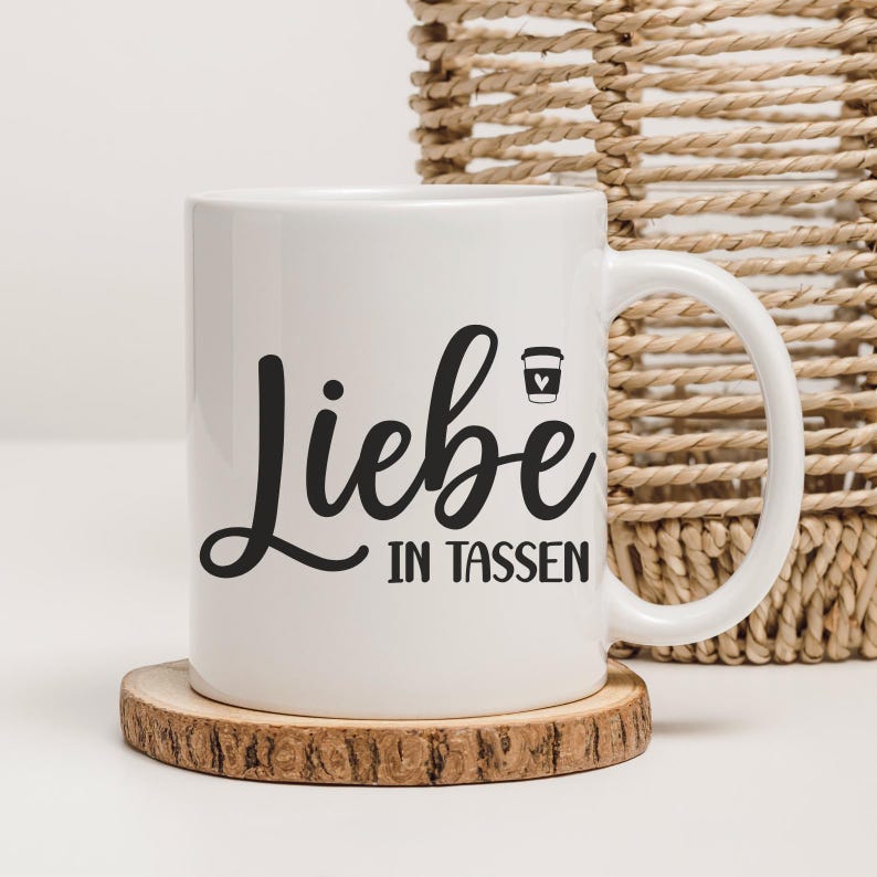 May include: White ceramic mug with the German phrase "Liebe in Tassen" in black script, meaning "Love in Cups." A small coffee cup icon is next to the text. The mug sits on a wooden coaster, with a woven basket in the background.