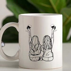 May include: White ceramic mug with a black line drawing of two women sitting back to back, raising a glass of wine in each hand. The women have long hair and are wearing casual clothing.