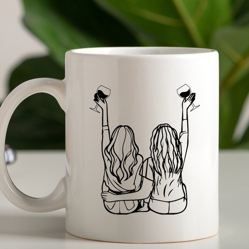 May include: White ceramic mug with a black line drawing of two women sitting back to back, raising a glass of wine in each hand. The women have long hair and are wearing casual clothing.