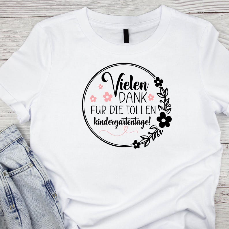 Peut inclure: T-shirt blanc avec un motif circulaire noir qui dit "Vielen Dank fur die tollen Kindergartentage!" avec des fleurs roses et des feuilles noires autour du cercle.