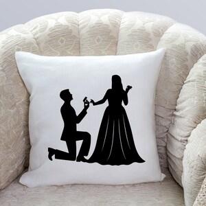 May include: A white pillow with a black silhouette of a couple getting engaged. The man is kneeling and holding a ring, while the woman is standing with her hand outstretched.