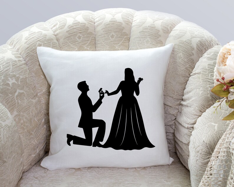May include: A white pillow with a black silhouette of a couple getting engaged. The man is kneeling and holding a ring, while the woman is standing with her hand outstretched.