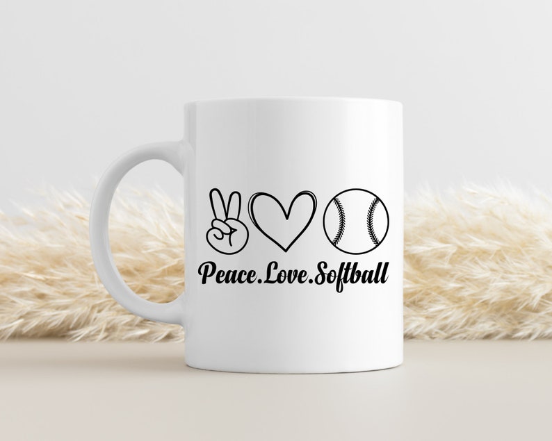 May include: White ceramic coffee mug with a black peace sign, heart, and softball graphic. The text "Peace. Love. Softball" is printed below the graphic.