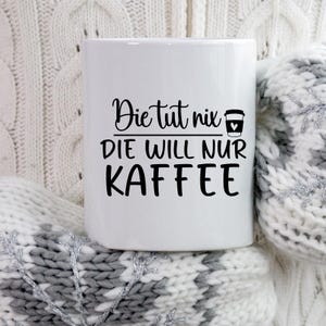 May include: White ceramic coffee mug with the text "Die tut nix, DIE WILL NUR KAFFEE" in black script. A coffee cup icon with a heart is next to the text. The mug is resting on a white and gray knitted surface.