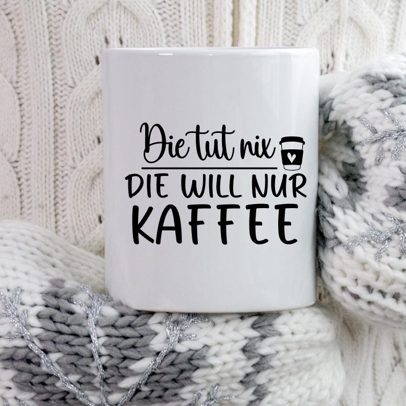 May include: White ceramic coffee mug with the text "Die tut nix, DIE WILL NUR KAFFEE" in black script. A coffee cup icon with a heart is next to the text. The mug is resting on a white and gray knitted surface.