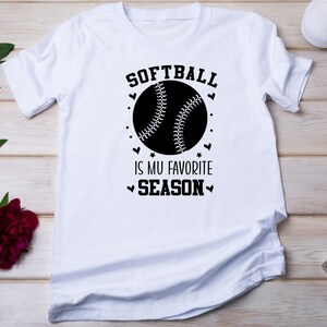 May include: White t-shirt with a black graphic of a softball and the text "Softball is my favorite season".