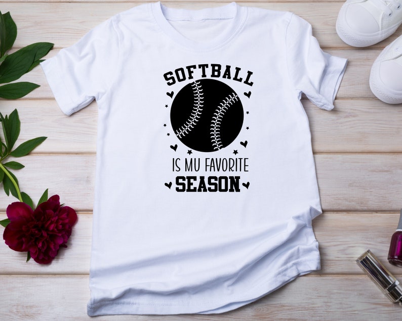 May include: White t-shirt with a black graphic of a softball and the text "Softball is my favorite season".