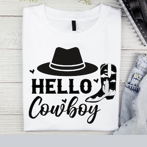 以下が含まれることがあります： 黒いカウボーイハットとブーツのグラフィックが描かれた白いTシャツ。グラフィックの下に「HELLO COWBOY」というテキストが印刷されています。