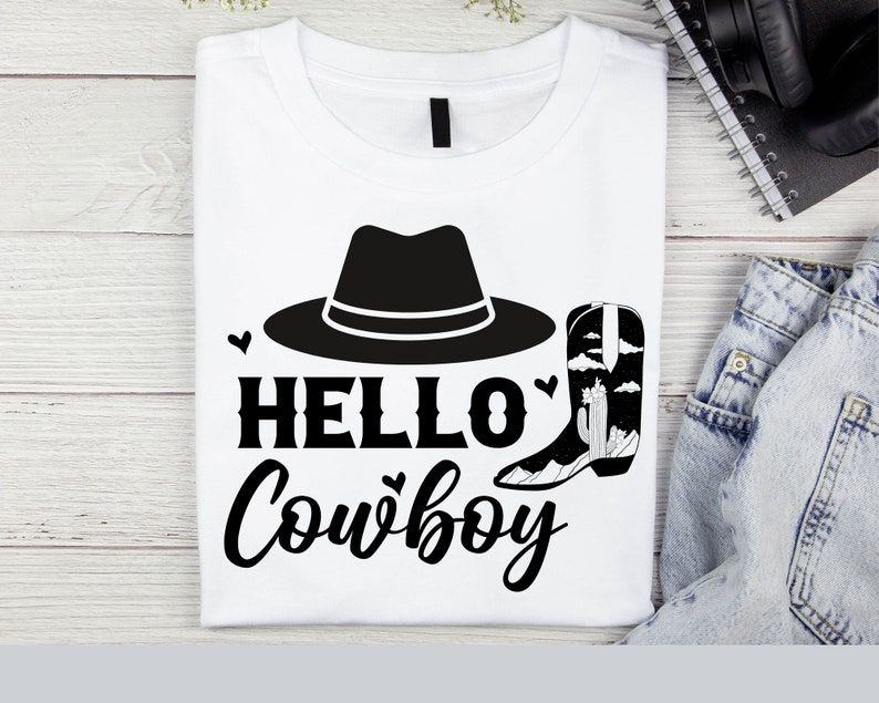 以下が含まれることがあります： 黒いカウボーイハットとブーツのグラフィックが描かれた白いTシャツ。グラフィックの下に「HELLO COWBOY」というテキストが印刷されています。