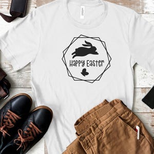 May include: White t-shirt with a black graphic of a bunny rabbit jumping with the text "Happy Easter" in a geometric frame.
