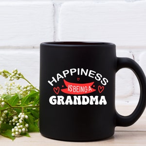 May include: Black ceramic coffee mug with white text that reads "Happiness is being a Grandma" with a red banner and three red hearts.