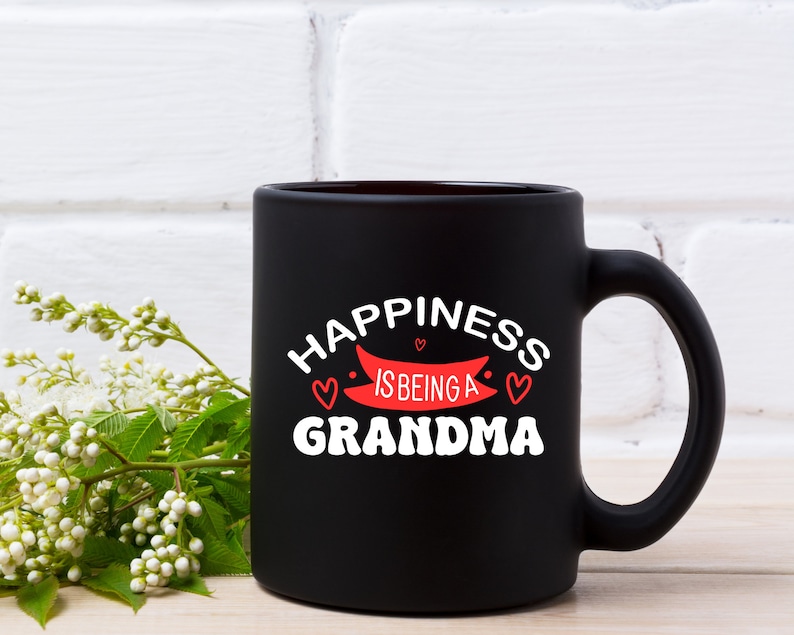 May include: Black ceramic coffee mug with white text that reads "Happiness is being a Grandma" with a red banner and three red hearts.