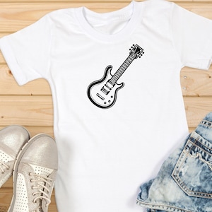 May include: White t-shirt with a black electric guitar graphic. The guitar design is centered on the shirt and is a detailed illustration. The shirt is paired with blue denim jeans and silver sneakers on a wooden surface.