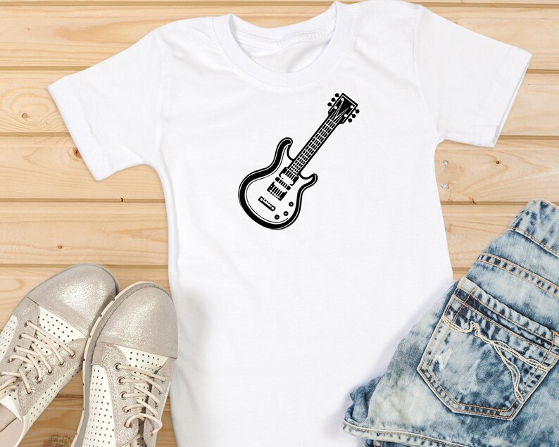 May include: White t-shirt with a black electric guitar graphic. The guitar design is centered on the shirt and is a detailed illustration. The shirt is paired with blue denim jeans and silver sneakers on a wooden surface.