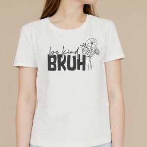 May include: White t-shirt with black text that reads "be kind BRUH" and a small floral design.