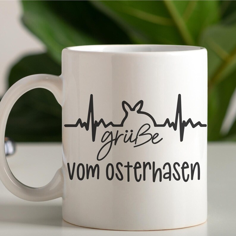 May include: White ceramic mug with a black graphic of a heartbeat with a bunny rabbit inside the beat. The text "gr&uuml;&szlig;e vom Osterhasen" is printed below the graphic.