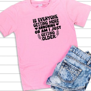 May include: Pink t-shirt with black text that reads "Is everyone getting more annoying or am I just getting older?" with a small heart and a leaf design.