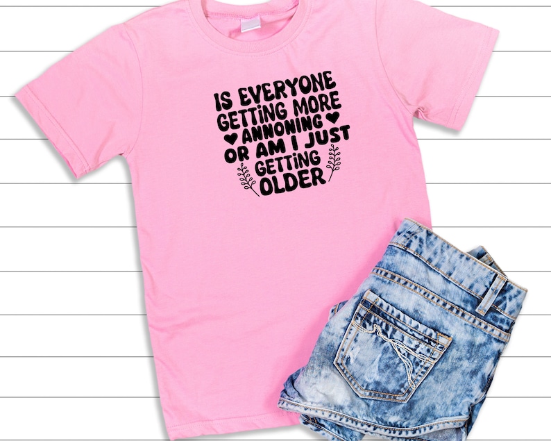 May include: Pink t-shirt with black text that reads "Is everyone getting more annoying or am I just getting older?" with a small heart and a leaf design.