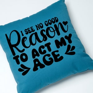 May include: A teal blue throw pillow with the text "I see no good reason to act my age" in black lettering with a small heart and leaf design.